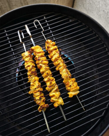 Drei goldbraun gegrillte Hähnchenspieße liegen über Flammen auf dem Rost eines Grills.