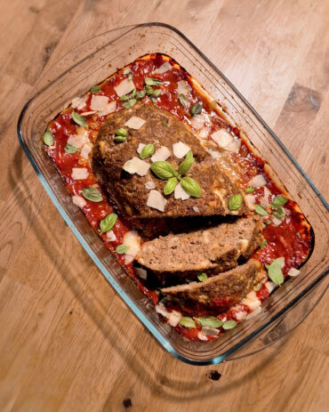 Ein aufgeschnittener Hackbraten liegt in Tomatensauce, garniert mit gehobeltem Parmesan und frischen Basilikumblättern.