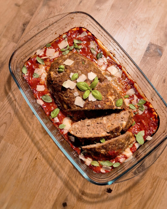 Ein aufgeschnittener Hackbraten liegt in Tomatensauce, garniert mit gehobeltem Parmesan und frischen Basilikumblättern.