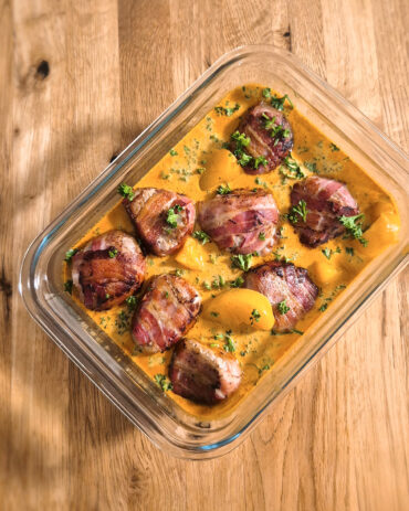 Im Ofen gegarte Schweinsmedaillons im Speckmantel liegen mit süssen Pfirsichstücken in cremiger Sauce mit feiner Currynote und Petersilie.